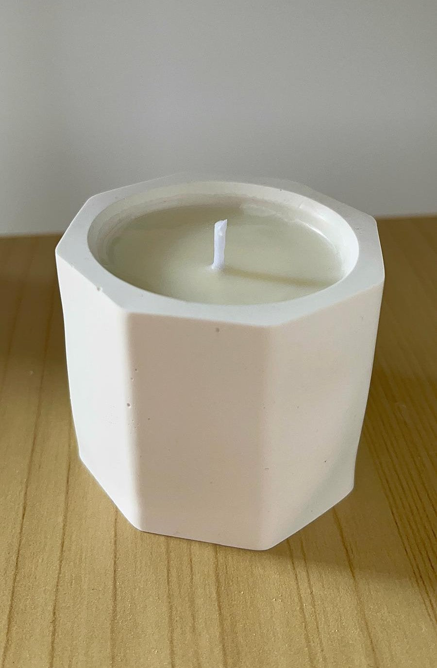 Vela de soja sin aroma en recipiente blanco de forma geométrica, colocada sobre superficie de madera clara