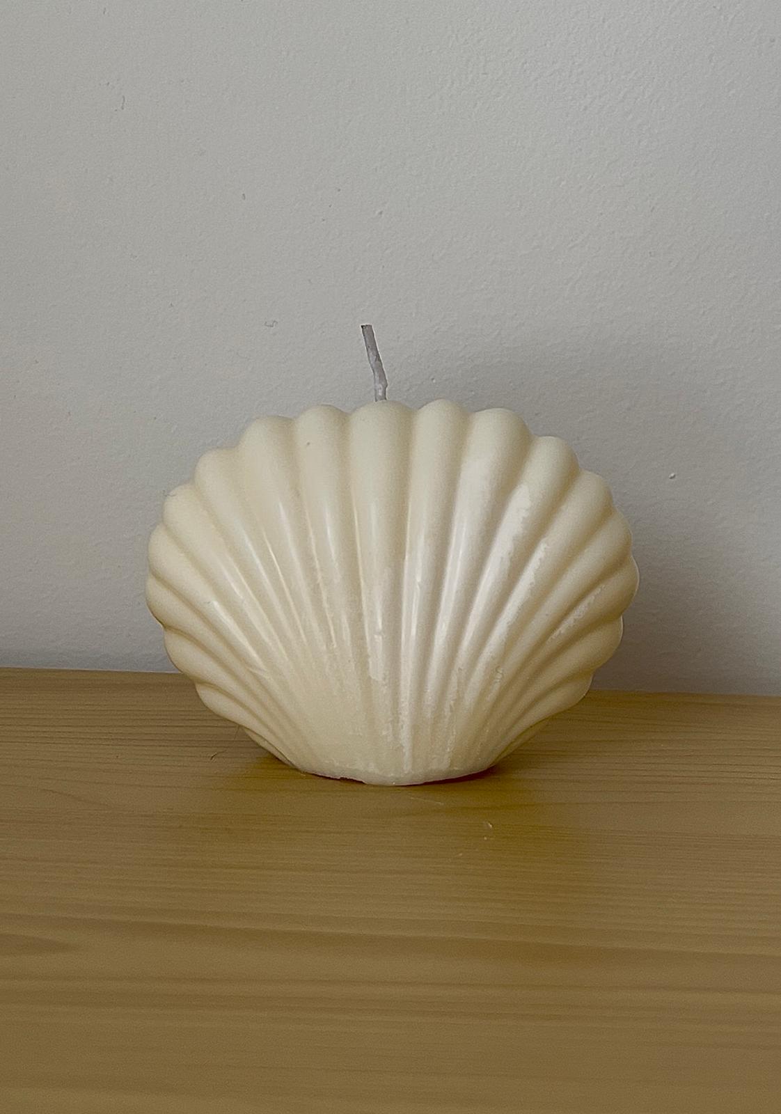 Vela decorativa con forma de concha de mar en tono marfil, colocada sobre superficie de madera clara.