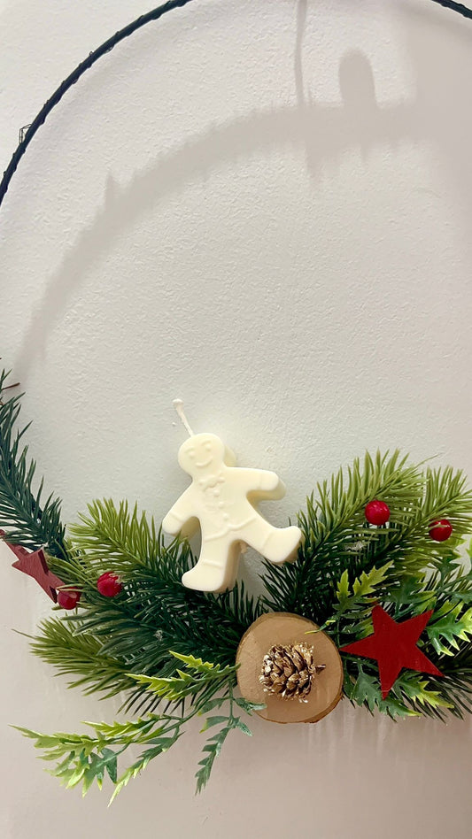 Vela decorativa Galleta de Invierno de Winged Essence en forma de muñeco de jengibre, hecha con cera de soja vegetal, sobre corona navideña con ramas y piñas.